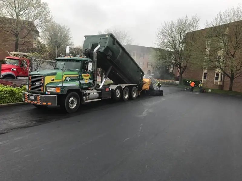 Industrial asphalt paving with dump truck feeding a paver at a warehouse yard